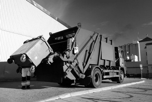 A waste collection vehicle with an open rear hopper, positioned on a paved surface next to a large industrial building with a corrugated metal wall, is in the process of collecting a standard large wheeled rubbish bin. The bin is made of plastic with a textured surface and has a lid on top, which is closed. Nearby, a worker in high-visibility trousers is standing close to the bin, operating the mechanical arm that lifts and empties the contents into the vehicle. The scene is outdoors, with a partly cloudy sky overhead, and the vehicle's black and white finish contrasts with the light-coloured pavement. This setting is likely within an industrial or commercial area in Kilburn, supporting the rubbish removal services provided by Office Clearance Kilburn, which specialises in waste collection and clearance services in the local area.