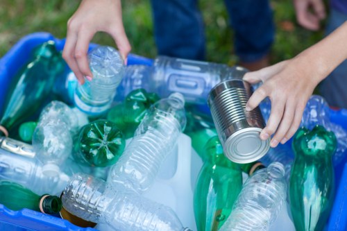 A person’s hands placing a clear plastic water bottle and a metal tin can into a light blue recycling bin filled with various discarded plastic bottles, including green and transparent ones, outdoors on a grassy area. The background shows part of the person's blue jeans and other hands reaching towards the bin, indicating a waste collection or recycling activity near Kilburn. The scene emphasizes waste separation and disposal, consistent with rubbish removal services offered by Office Clearance Kilburn, serving the Kilburn area in northwest London. The clear plastic bottles are crumpled, and the tin can has a metallic finish, contrasting with the smooth plastic surfaces, all situated on a paved or grassy ground, with natural daylight illuminating the scene.