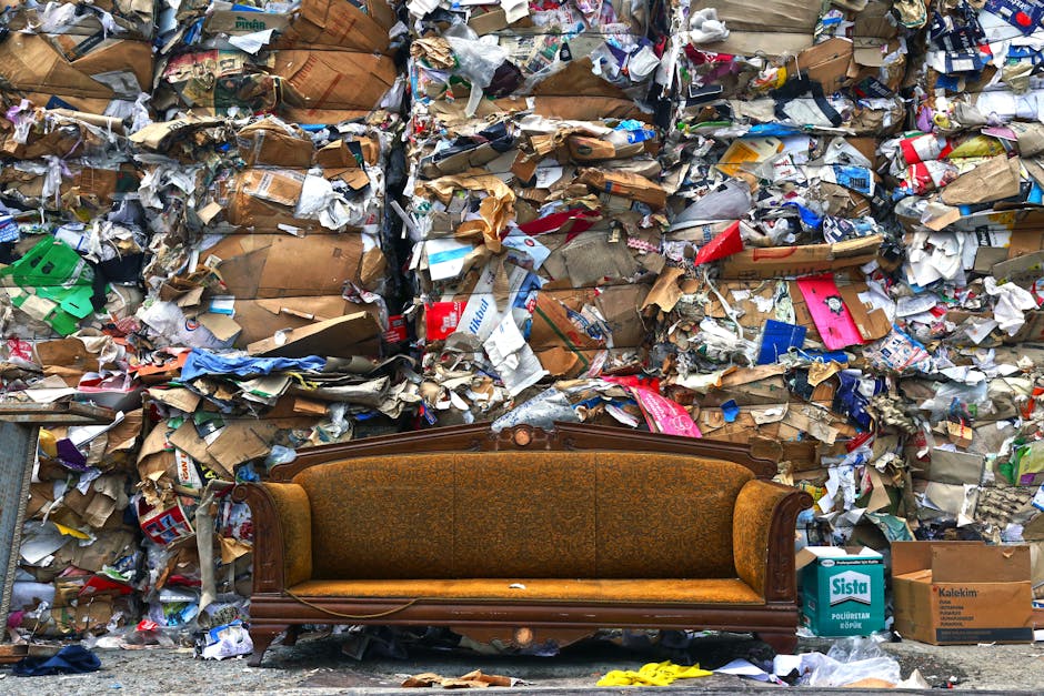 A vintage brown upholstered sofa with wooden frame and armrests positioned in front of a large, densely packed wall of mixed waste and recyclable materials, including crumpled cardboard boxes, shredde