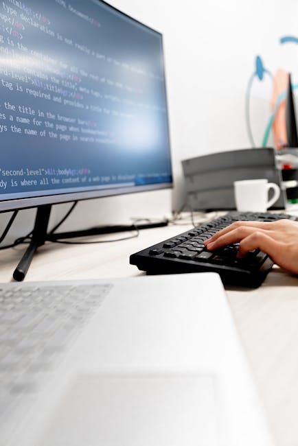 A person seated at a desk, viewed from behind, working on a laptop that displays a screen filled with colorful lines of programming code. The individual is wearing a light blue shirt and holds a pen i