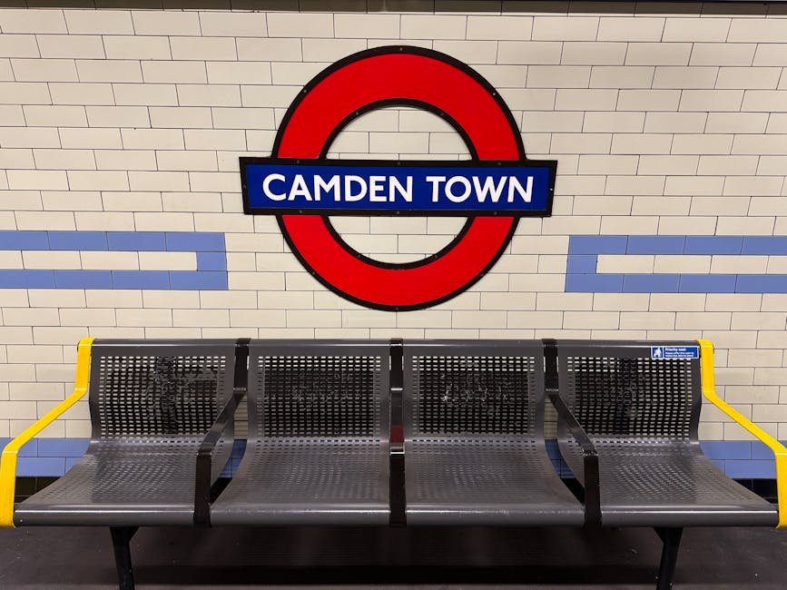 A row of four metal bench seats with perforated surfaces and curved backs is situated on a tiled floor in a subway station, directly beneath a large circular London Underground sign featuring a red ri