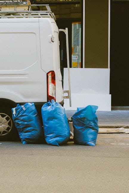 Two large black plastic bags filled with waste materials are placed on a sidewalk in front of a black metal fence and a hedge with dense foliage. The bags appear to contain household or garden rubbish